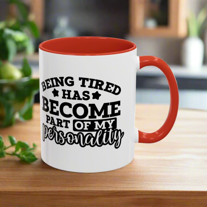 White coffee mug with red handle and rim featuring black text that reads ’Being tired has become part of my personality’