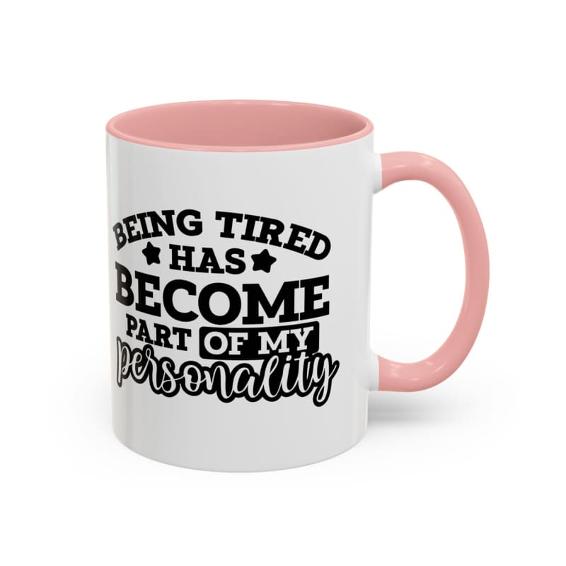 White coffee mug with pink handle and rim featuring black text that reads ’Being tired has become part of my personality’