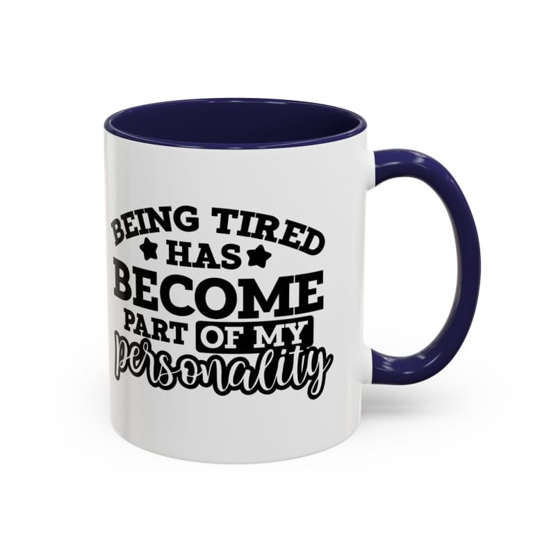 White coffee mug with navy blue handle and interior featuring black text that reads ’Being tired has become part of my personality’