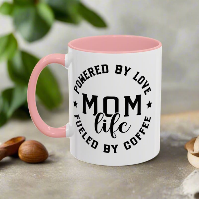 White coffee mug with pink handle featuring ’MOM LIFE’ text and ’Powered by Love, Fueled by Coffee’ message.